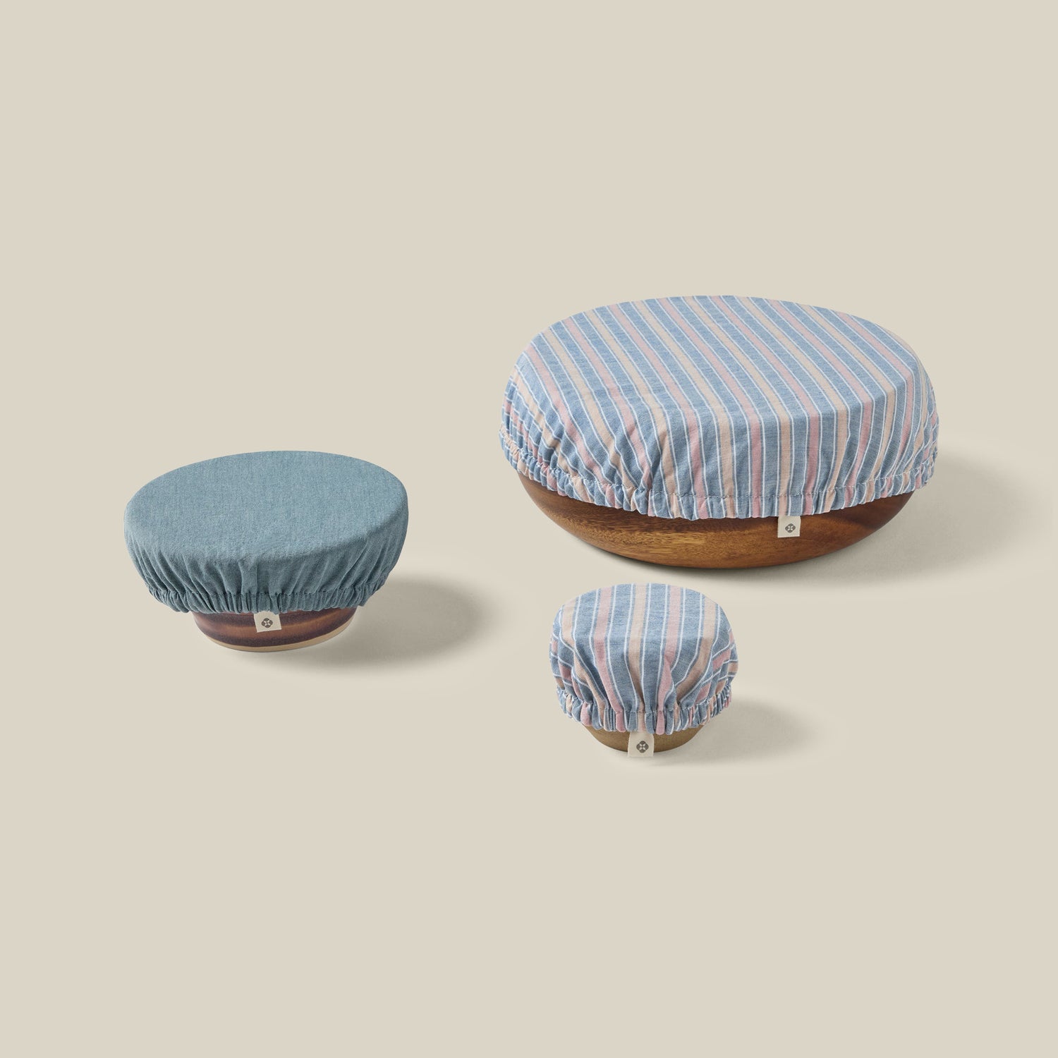 Three fabric stretch covers in blue, light blue, and pink striped patterns, shown stretched over round wooden bases on a solid beige background.- Number 1