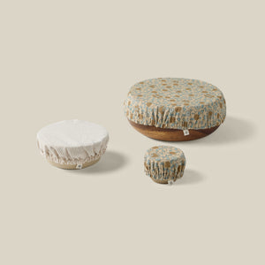 Three round fabric food storage covers in different sizes, displayed on top of wooden bowls against a solid beige background. The covers feature a floral pattern in muted blue, brown, and cream, with one cover on the left being a solid cream ribbed fabric. - Number 1