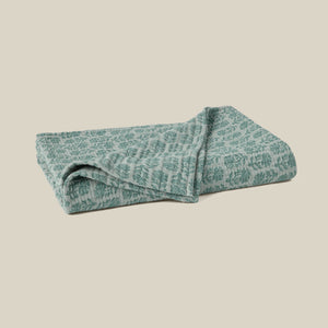 A folded quilted blanket with a sage green floral block print on a white background, set against a solid beige backdrop. - Number 1