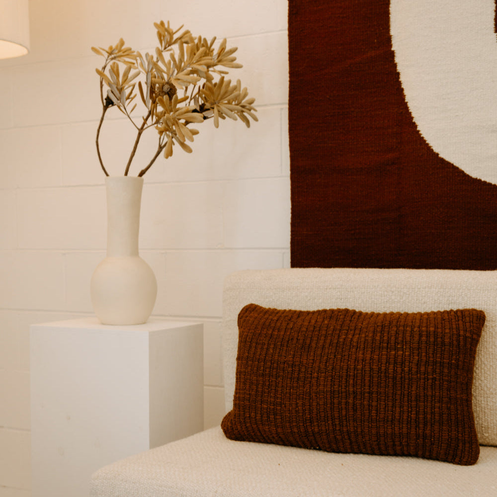 Brown textured pillow on a beige sofa with a vase of dried plants on a white side table.- Number 2