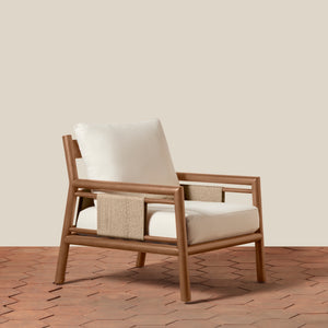 A modern armchair with a light brown wood frame, cream-colored cushions, and a woven beige accent panel on the side, set against a solid beige wall and terracotta tile floor. - Number 1