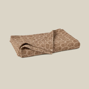 A folded quilted blanket in a warm taupe-brown color with a lighter beige leafy pattern, set against a solid light beige background. - Number 1