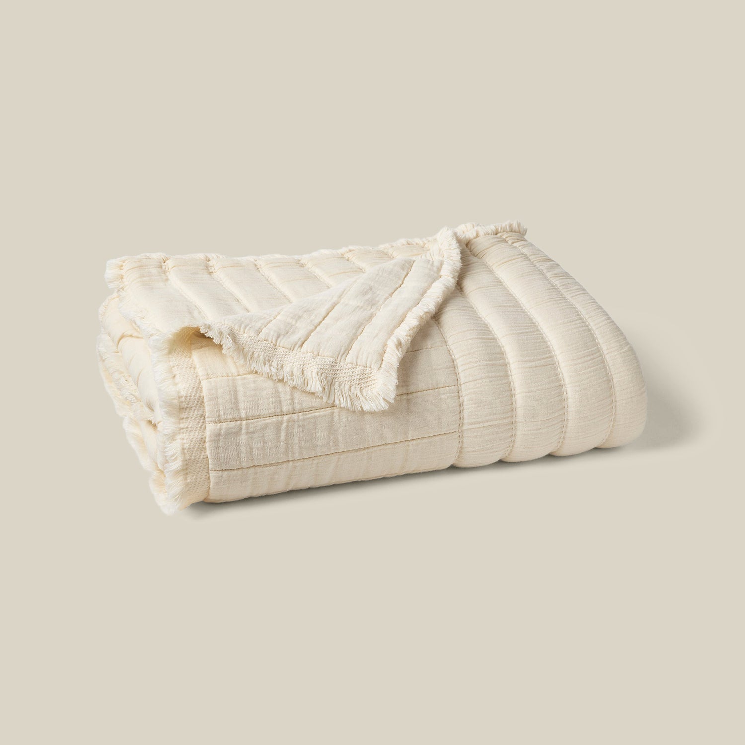 A folded cream-colored textured blanket with a ribbed pattern and fringed edges, displayed on a solid light beige background.- Number 1