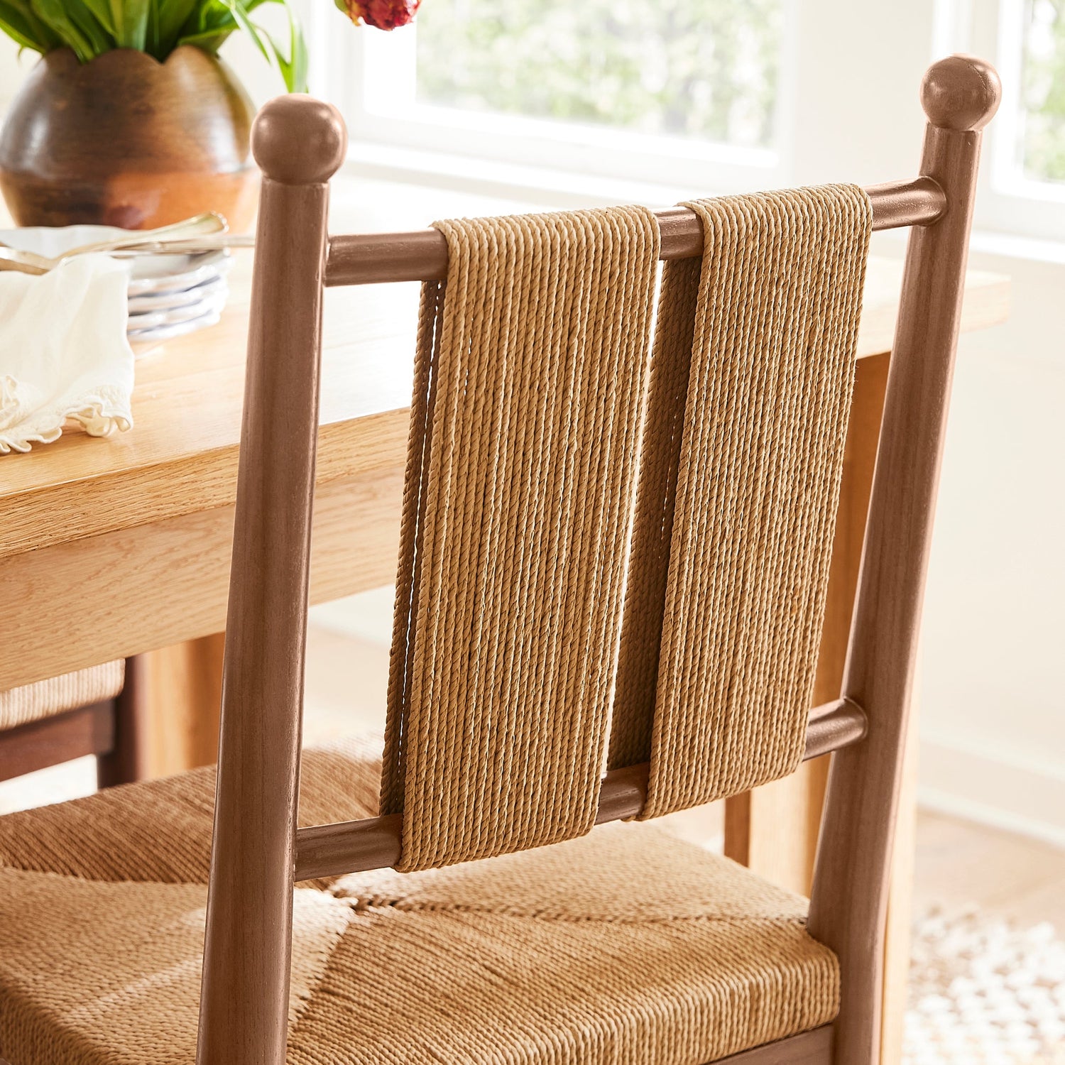 A wooden dining chair with woven natural-fiber seat and back slats, shown in a bright dining room near a window with tulips in the background.- Number 3