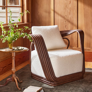 A modern accent chair with a dark brown curved frame and plush cream bouclé seat and back cushion, styled in a warm wood-paneled room next to a small side table with a plant. - Number 2