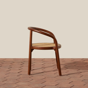 A mid-century modern wooden armchair with a curved bentwood frame and a woven cane seat, shown on a terracotta hexagonal tile floor against a solid beige background. - Number 8