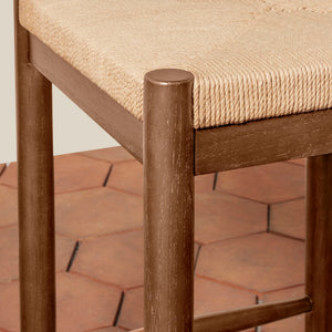 Close-up of a wooden stool with a woven beige rope seat and dark brown legs, shown in a real interior setting with terracotta tile flooring. - Number 7