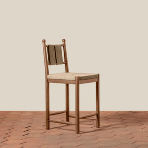 A wooden bar stool with a woven rope backrest and a light beige upholstered seat, set against a solid beige wall and a brown hexagonal tile floor. - Number 1