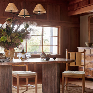 A rustic dining room scene with a round wooden table, four matching wooden chairs with woven seats, a large floral arrangement in a vase, and a chandelier above. The room has wood-paneled walls and a window in the background. - Number 11