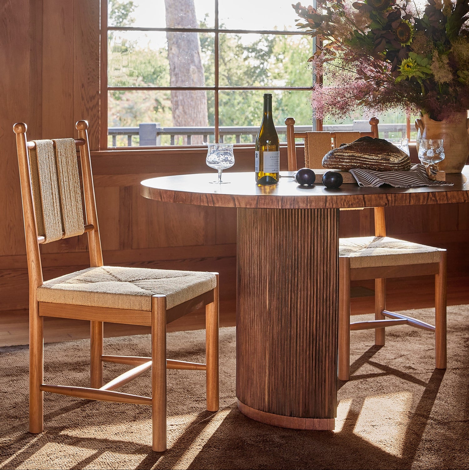 A rustic dining room scene featuring a round wooden table with a fluted pedestal base and two matching wooden chairs with woven seat and back cushions, set in front of a large window with natural light.- Number 10