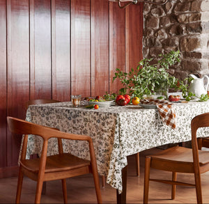A rustic dining room scene with a floral tablecloth on a long table, surrounded by wooden chairs. The table is set with greenery, tomatoes, and a white pitcher, against a backdrop of dark wood paneling and a stone wall. - Number 2