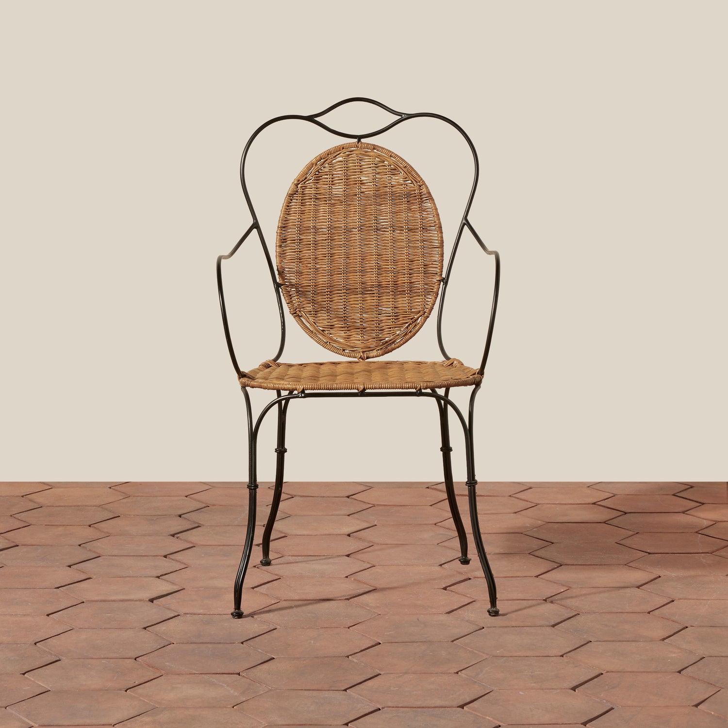 A vintage-style metal and wicker dining chair with a curved black wrought-iron frame, a round woven rattan backrest, and a woven seat, set against a beige wall and terracotta tile floor.- Number 5