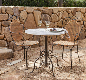 Outdoor patio dining set featuring a round white marble-top table with a black metal pedestal base, accompanied by two wicker-back and wicker-seat chairs with black metal frames, set on a stone patio in front of a rustic stone wall. - Number 2