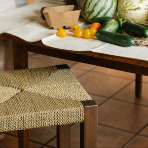 A rustic table setup with fresh vegetables including a watermelon, cucumbers, and small yellow tomatoes, alongside woven baskets, paper bags, and bottles in the background. - Number 3