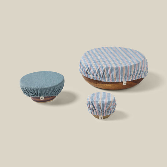 Three fabric stretch covers in blue, light blue, and pink striped patterns, shown stretched over round wooden bases on a solid beige background.