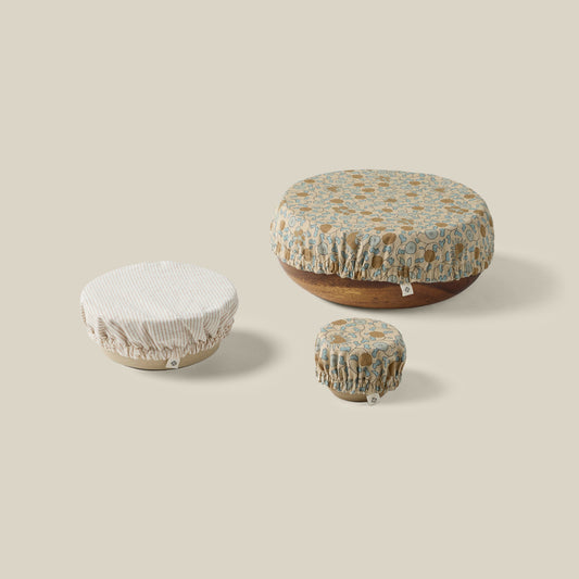 Three round fabric food storage covers in different sizes, displayed on top of wooden bowls against a solid beige background. The covers feature a floral pattern in muted blue, brown, and cream, with one cover on the left being a solid cream ribbed fabric.