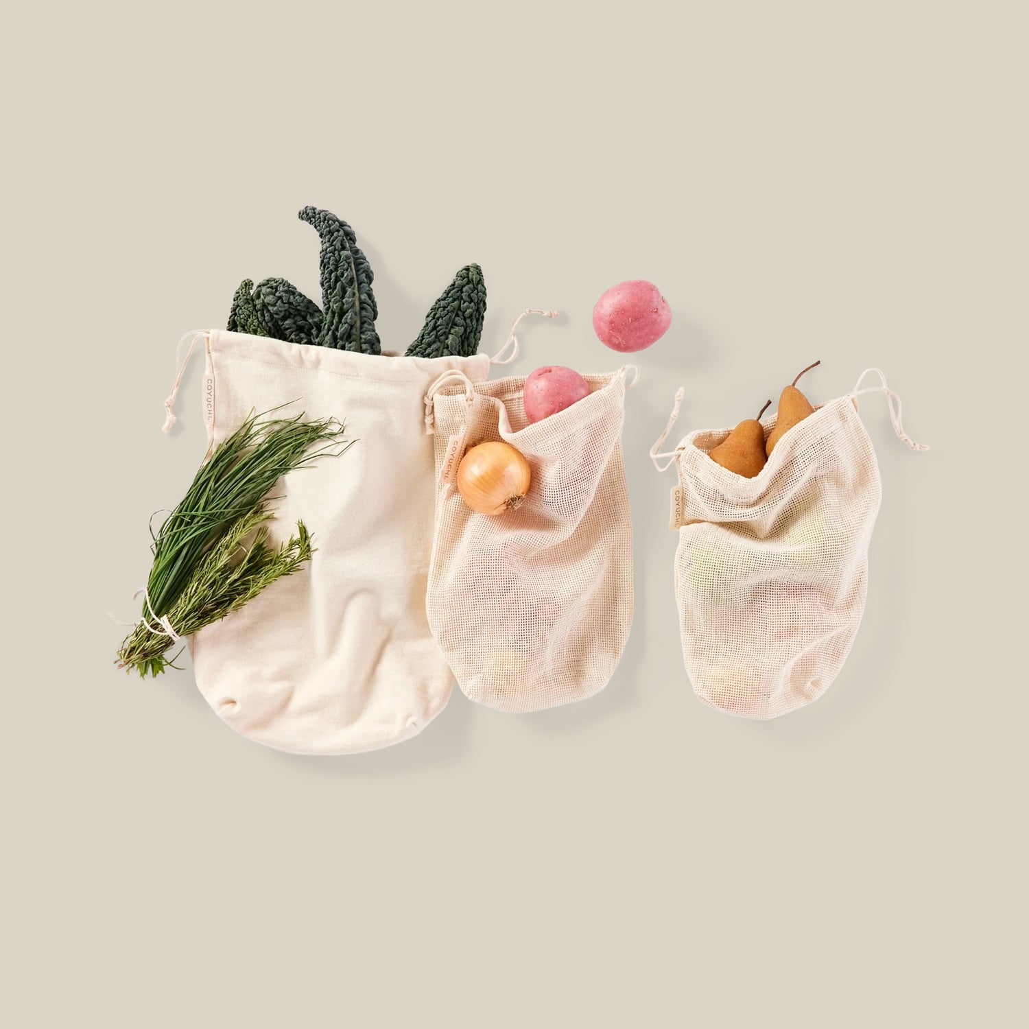 Three natural canvas drawstring mesh bags arranged on a light beige background, filled with produce including a dark leafy vegetable, a pink onion, a yellow onion, and a pear.- Number 1