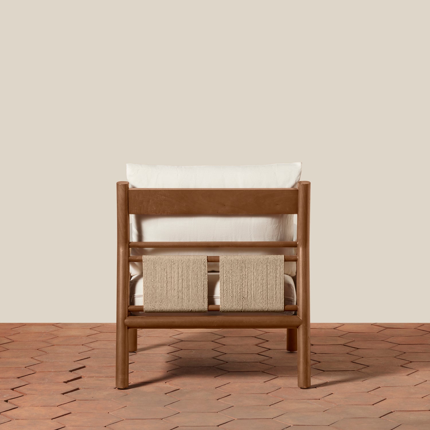 A wooden lounge chair with a white cushion and a woven beige seat strap, shown from the back against a solid beige wall and terracotta hexagonal tile floor.- Number 5