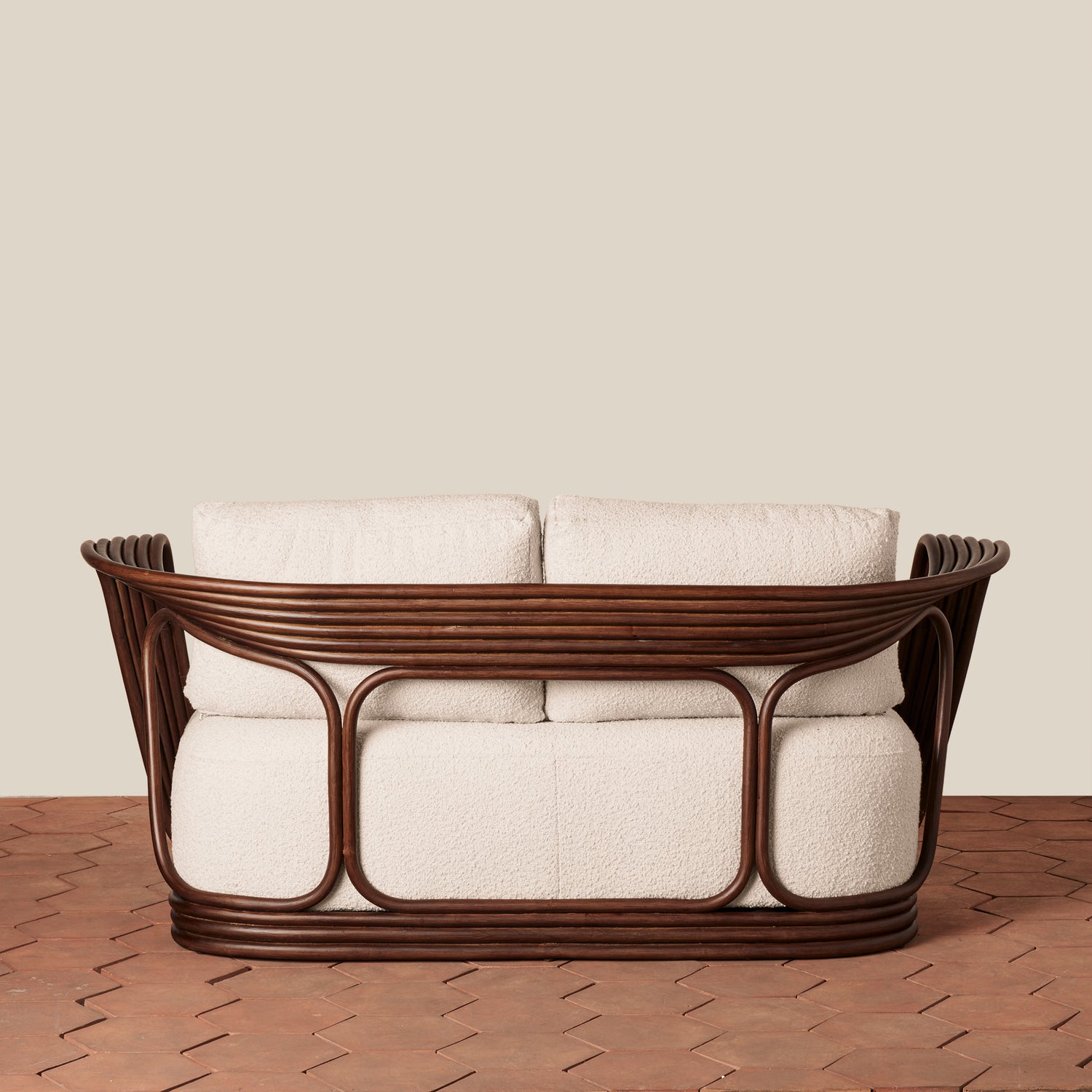 A modern loveseat with a curved dark brown woven rattan frame and thick cream cushions, set against a solid beige wall and a brown woven floor.- Number 4