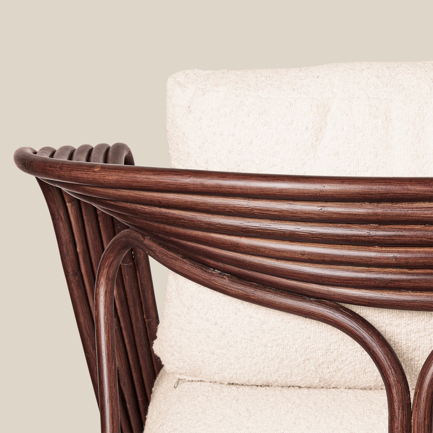 Close-up of a rattan armchair with a curved dark brown frame and a thick cream-colored textured cushion, set against a solid light beige background.- Number 6