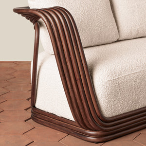 A close-up of a mid-century modern sofa corner featuring a curved, ribbed dark brown rattan or wicker frame with cream textured upholstery, set on a terracotta tile floor. - Number 7