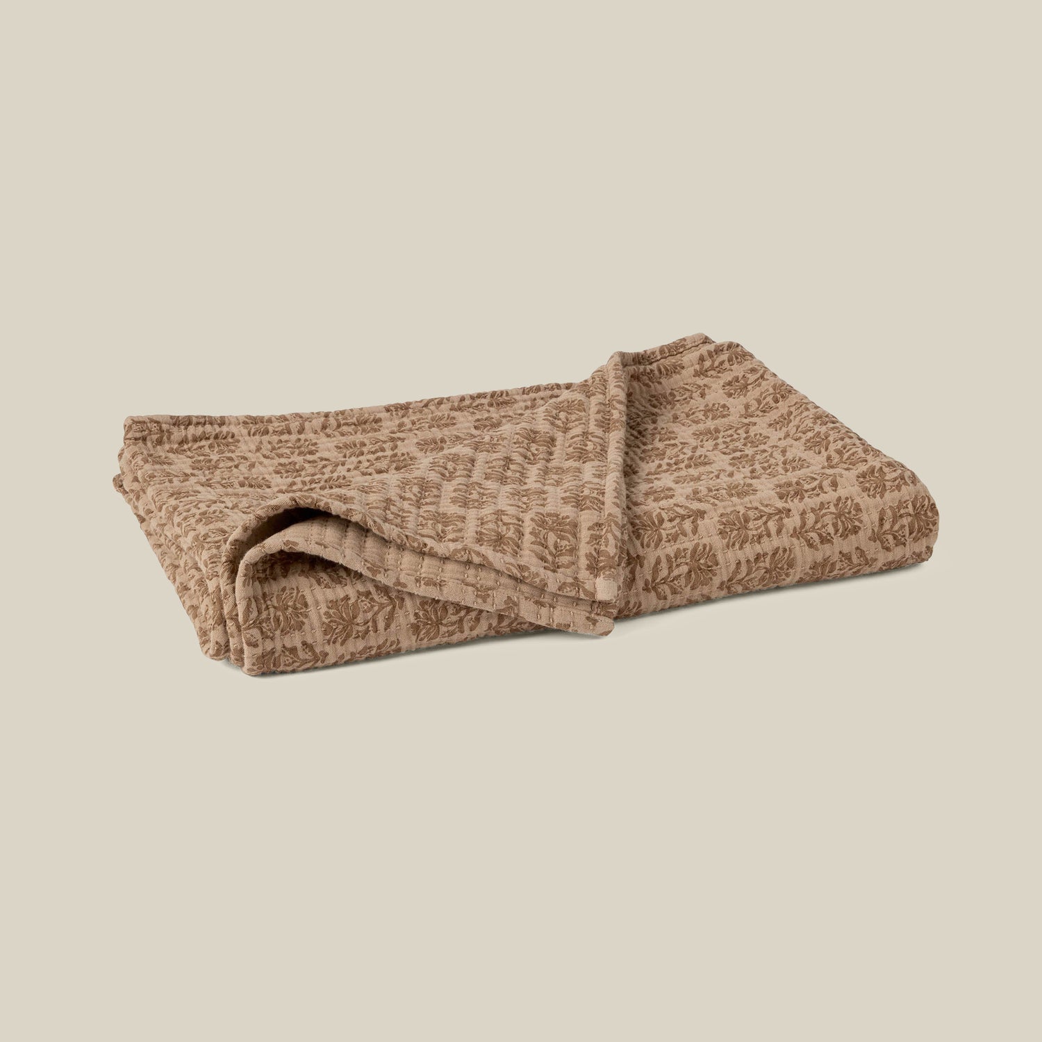 A folded quilted blanket in a warm taupe-brown color with a lighter beige leafy pattern, set against a solid light beige background.- Number 1