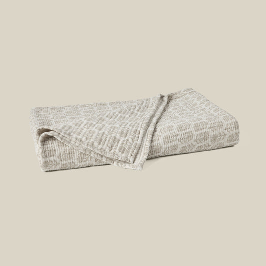 A folded textured blanket in a light beige and gray heathered pattern, set against a solid beige background.