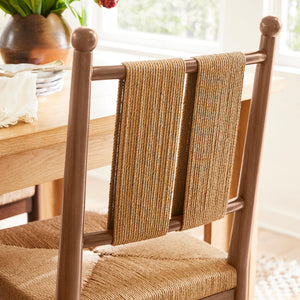 A wooden dining chair with woven natural-fiber seat and back slats, shown in a bright dining room near a window with tulips in the background. - Number 3