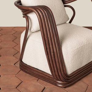 A close-up of a modern armchair with a curved dark brown rattan frame and thick, textured cream-colored cushions, set on a terracotta hexagonal tile floor against a neutral beige wall. - Number 7