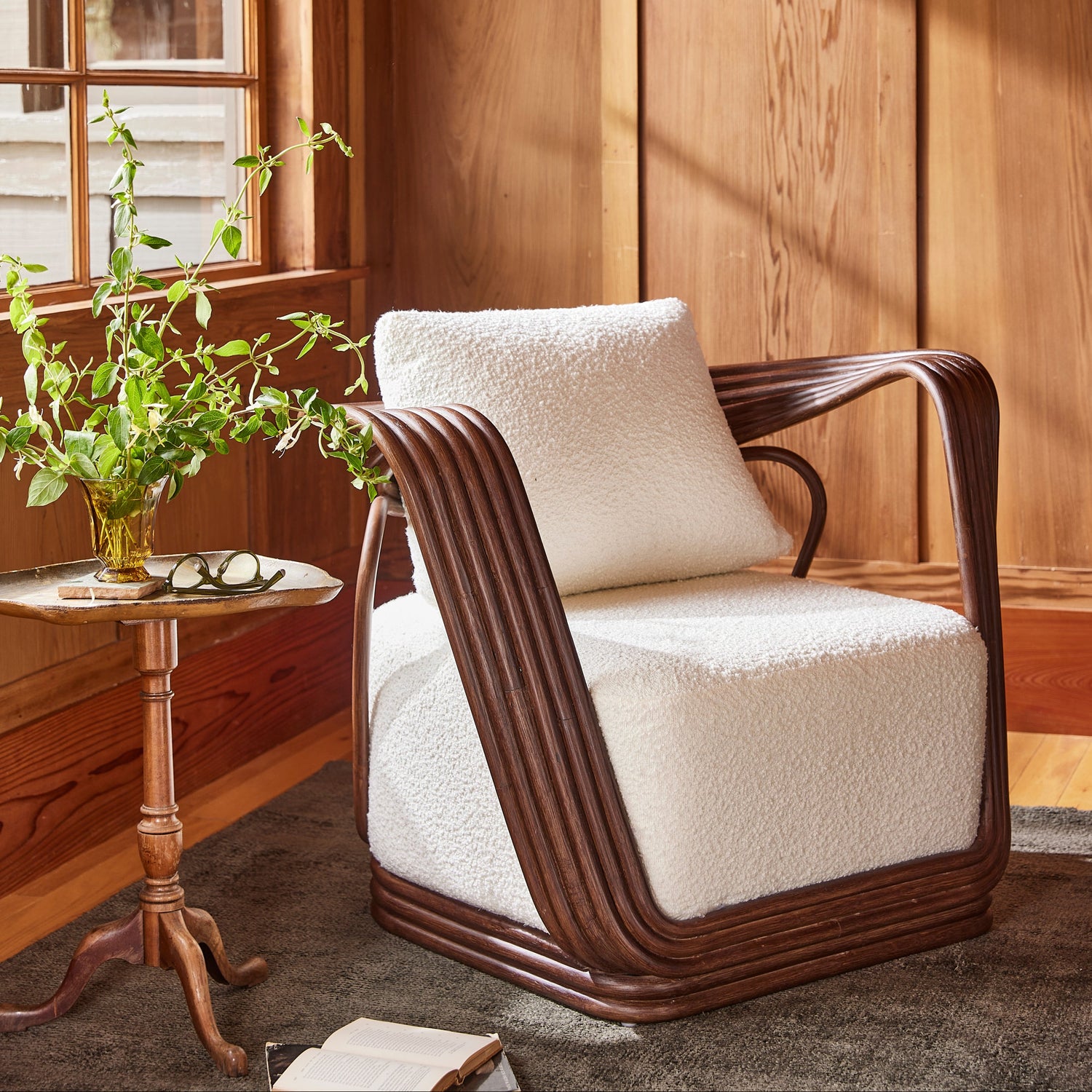A modern accent chair with a dark brown curved frame and plush cream bouclé seat and back cushion, styled in a warm wood-paneled room next to a small side table with a plant.- Number 2