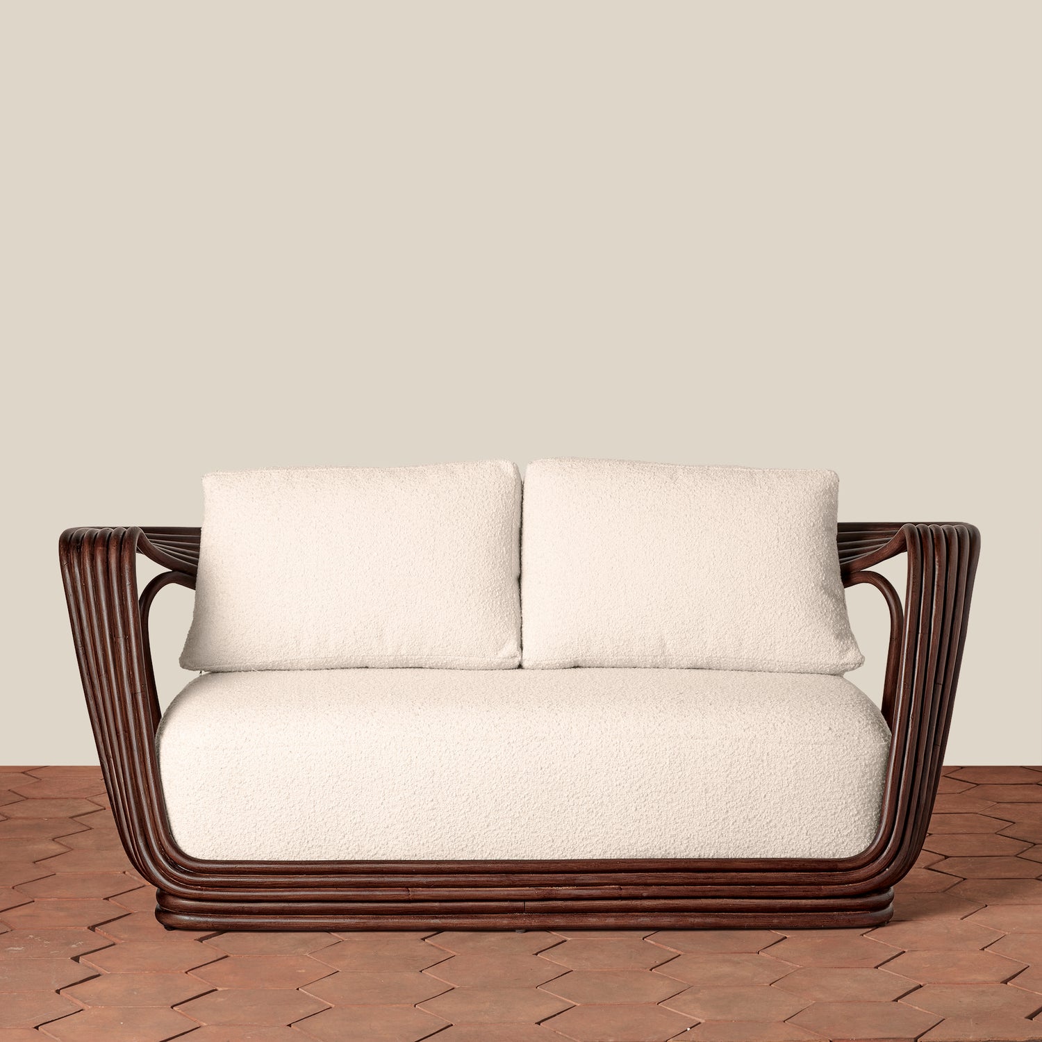 A modern loveseat with a dark brown curved wicker frame and thick off-white cushions, set against a plain beige wall and a brown brick floor.- Number 3