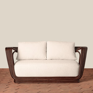 A modern loveseat with a dark brown curved wicker frame and thick off-white cushions, set against a plain beige wall and a brown brick floor. - Number 3