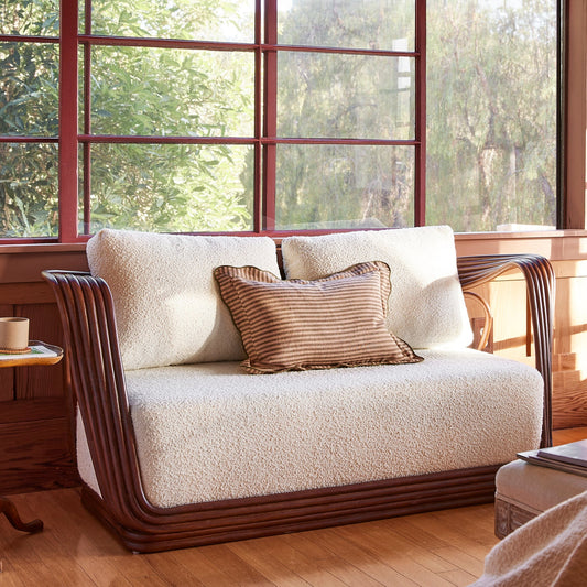 A cream loveseat with dark wood arms and legs, styled in a sunlit room with red-framed windows and a small wooden side table beside it. - Image 2