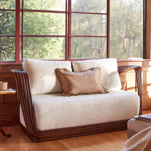 A cream loveseat with dark wood arms and legs, styled in a sunlit room with red-framed windows and a small wooden side table beside it. - Number 2