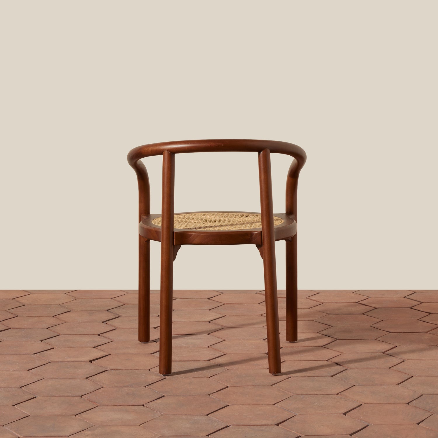 A wooden dining chair with a curved backrest and woven cane seat, shown from the front on a terracotta hexagonal tile floor against a solid beige background.- Number 10