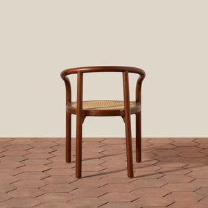 A wooden dining chair with a curved backrest and woven cane seat, shown from the front on a terracotta hexagonal tile floor against a solid beige background. - Number 10
