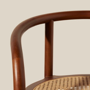 Close-up detail of a wooden chair with a curved medium-brown frame and a natural woven cane seat against a solid light beige background. - Number 11