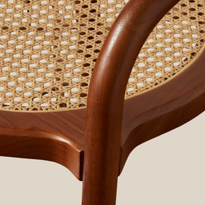 Close-up detail of a wooden chair with a curved medium-brown wood frame and a natural woven cane seat, set against a solid light beige background. - Number 12