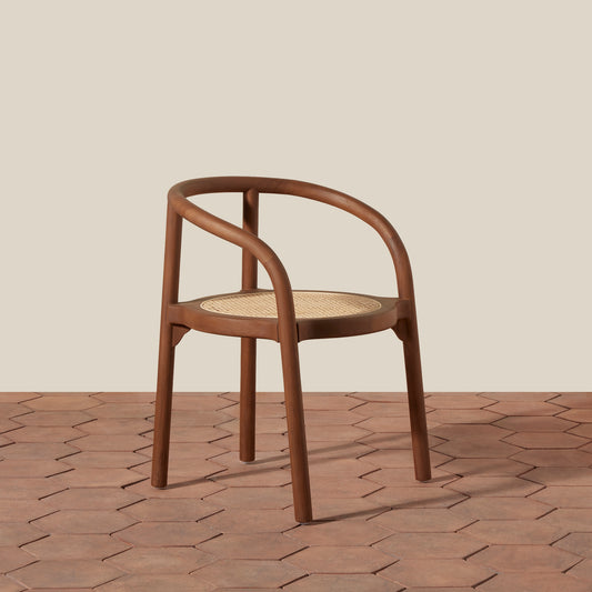 A mid-century modern wooden dining chair with a curved armrest, brown wood frame, and a woven cane seat, set against a beige wall and terracotta hexagonal tile floor.