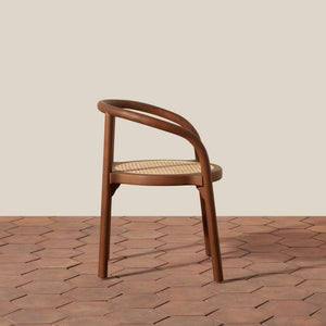 A mid-century modern wooden armchair with a curved backrest and a woven cane seat, positioned on a terracotta hexagonal tile floor against a solid beige background. - Number 2