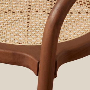 Close-up detail of a modern bistro chair featuring a curved medium-brown wooden frame and a natural woven cane seat, set against a solid light beige background. - Number 6