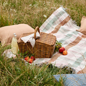A wicker picnic basket with a white cloth tied to it, placed on a plaid blanket in a grassy outdoor setting with pillows and fruit nearby. - Number 2