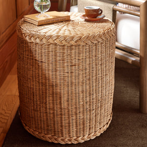 A round woven rattan side table or stool in a warm natural tan color, styled in a cozy room setting with a small glass vase of white flowers and a cup of coffee on top, next to a wooden chair with white cushions. - Number 2