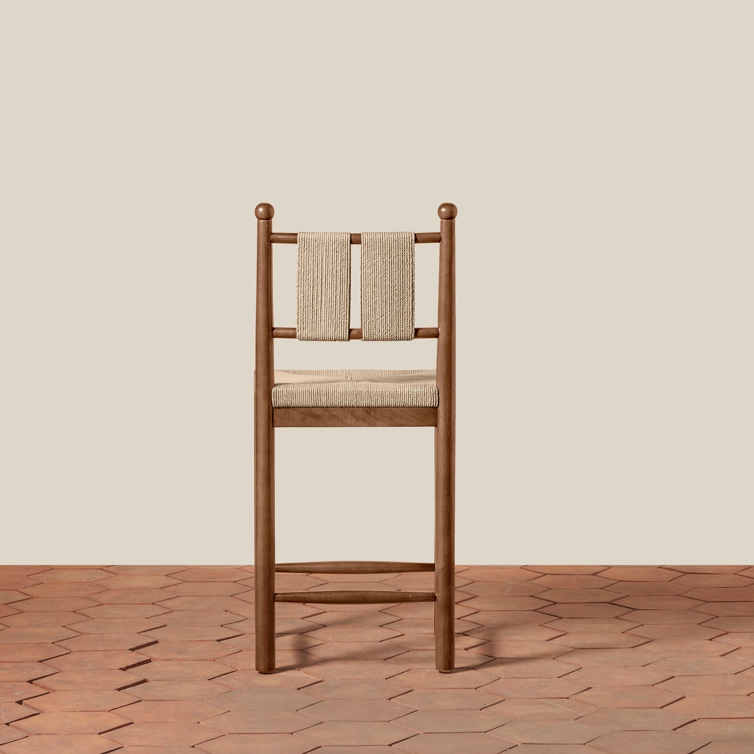 A wooden barstool with a woven beige seat and backrest, set against a solid beige wall and a terracotta hexagonal tile floor.- Number 5
