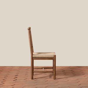 A single wooden dining chair with a woven beige seat cushion, shown from the side against a plain beige wall and terracotta hexagonal tile floor. - Number 12