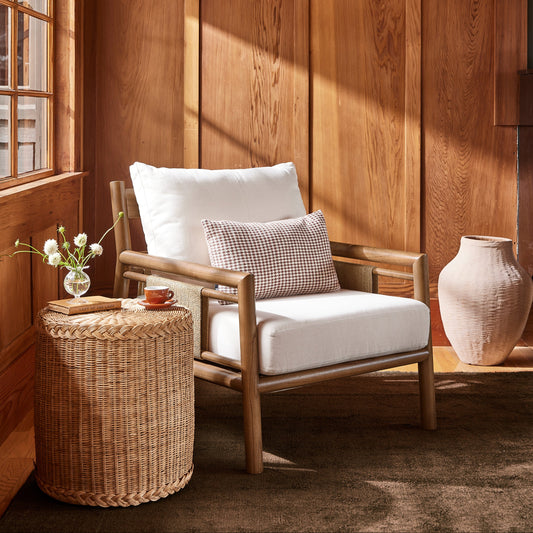 A wooden armchair with white cushions and a small patterned accent pillow, styled in a warm wood-paneled room with a woven side table, vase, and window light. - Image 2