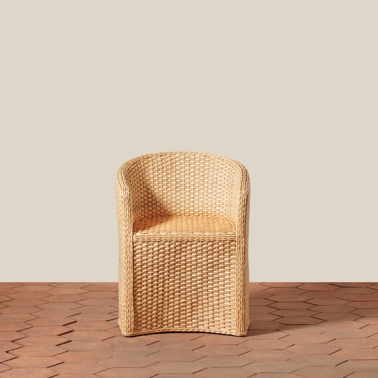 A single woven armchair in a natural tan wicker or rattan material, shown against a plain warm beige wall and a brown hexagonal tile floor.- Number 5
