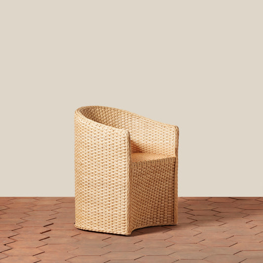 A single woven armchair in a natural tan rattan or wicker material, shown on a wood-look floor against a plain beige wall.