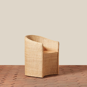 A single woven armchair in a natural tan rattan or wicker material, shown on a wood-look floor against a plain beige wall. - Number 1