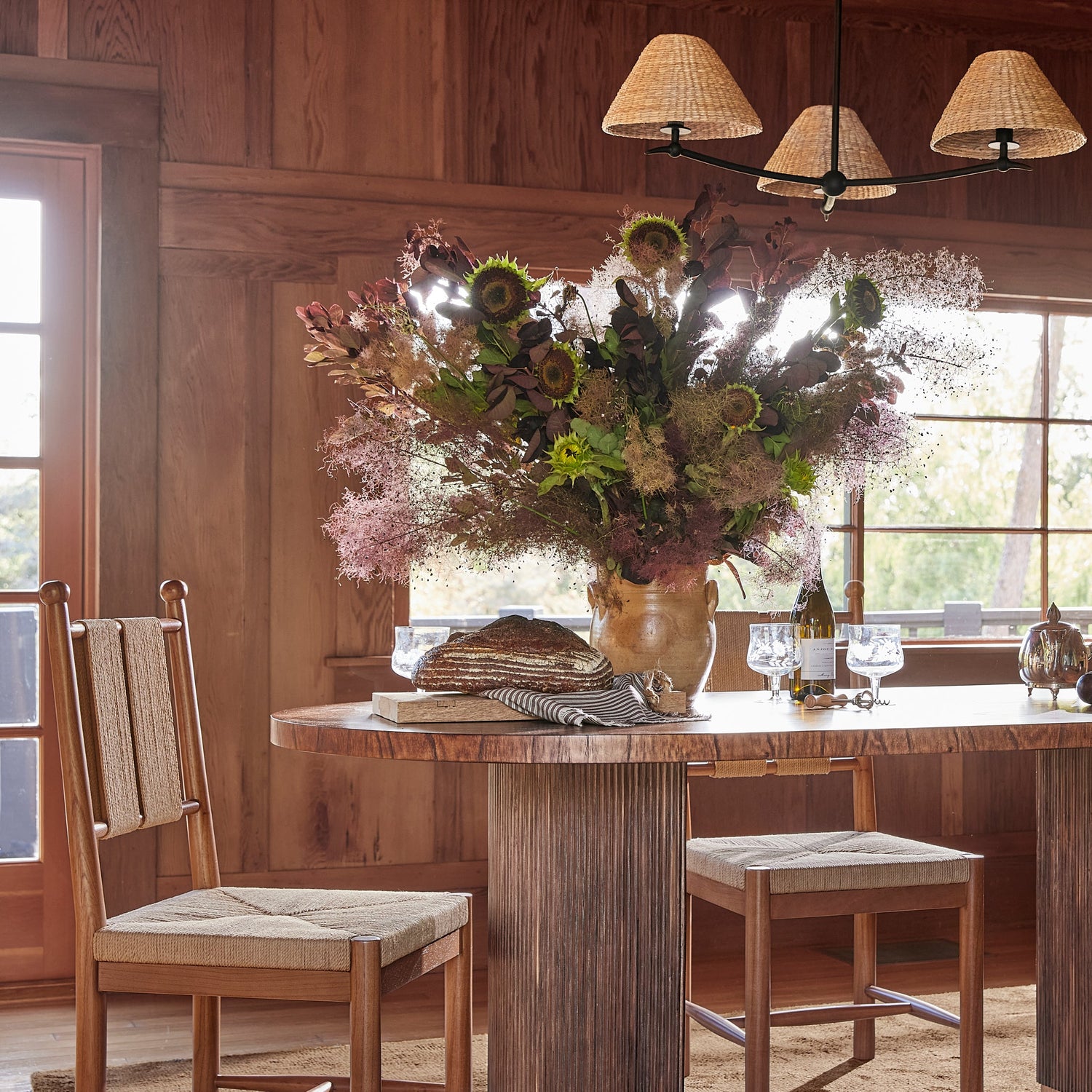 A rustic dining room scene with a large floral arrangement in a vase on a wooden table, with two wooden chairs and stools, a hanging lamp, and wood-paneled walls in the background.- Number 2